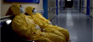 Two figures in bright yellow full-body PPE slump on a hospital bench.