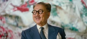 Michael Chow wears a blue suit with a tie and pocket square and oversized round glasses. He is looking to the right of the camera and smiling.