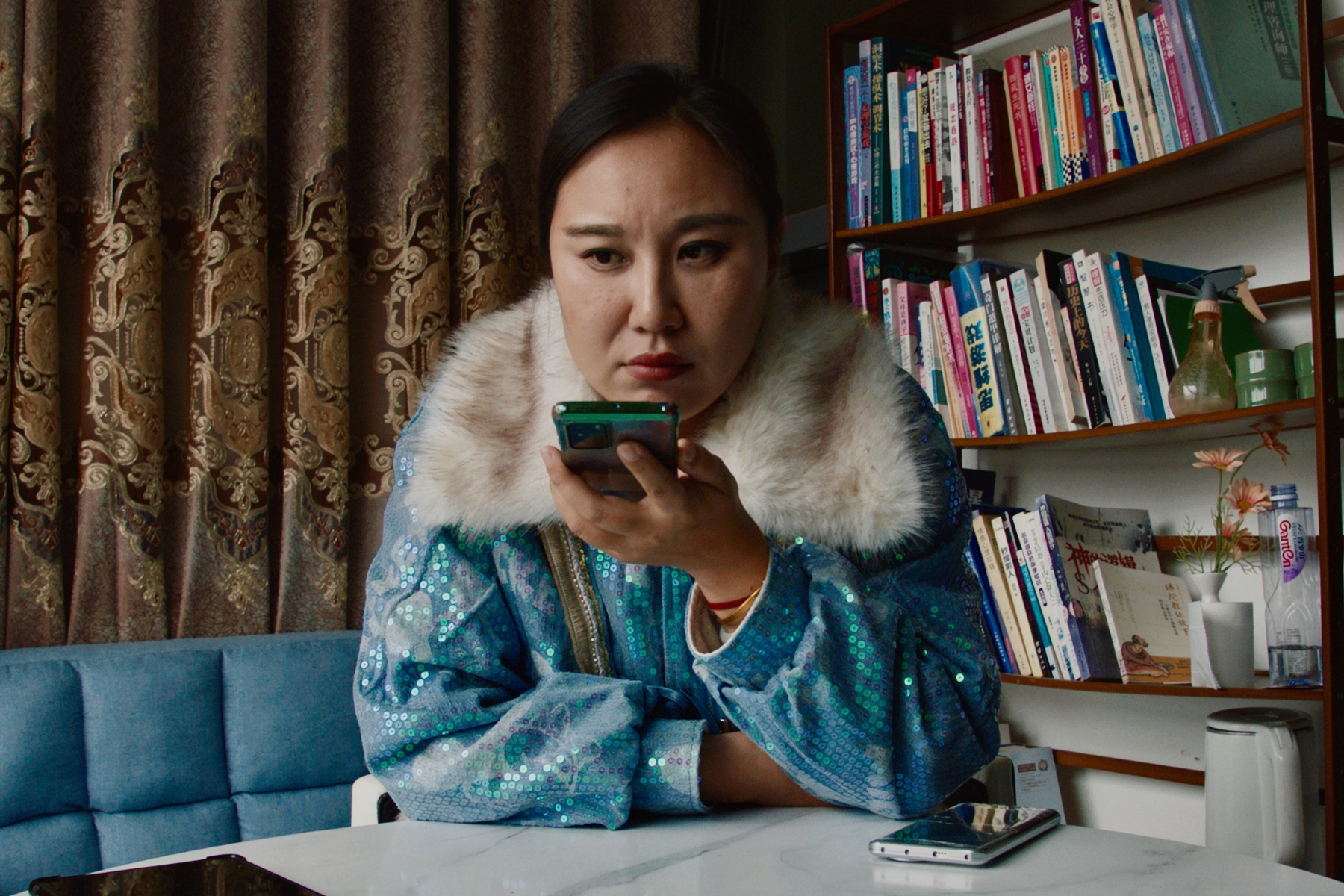 A person in a sparkly blue jacket with a faux fur collar speaks into a mobile phone held our in front of them.