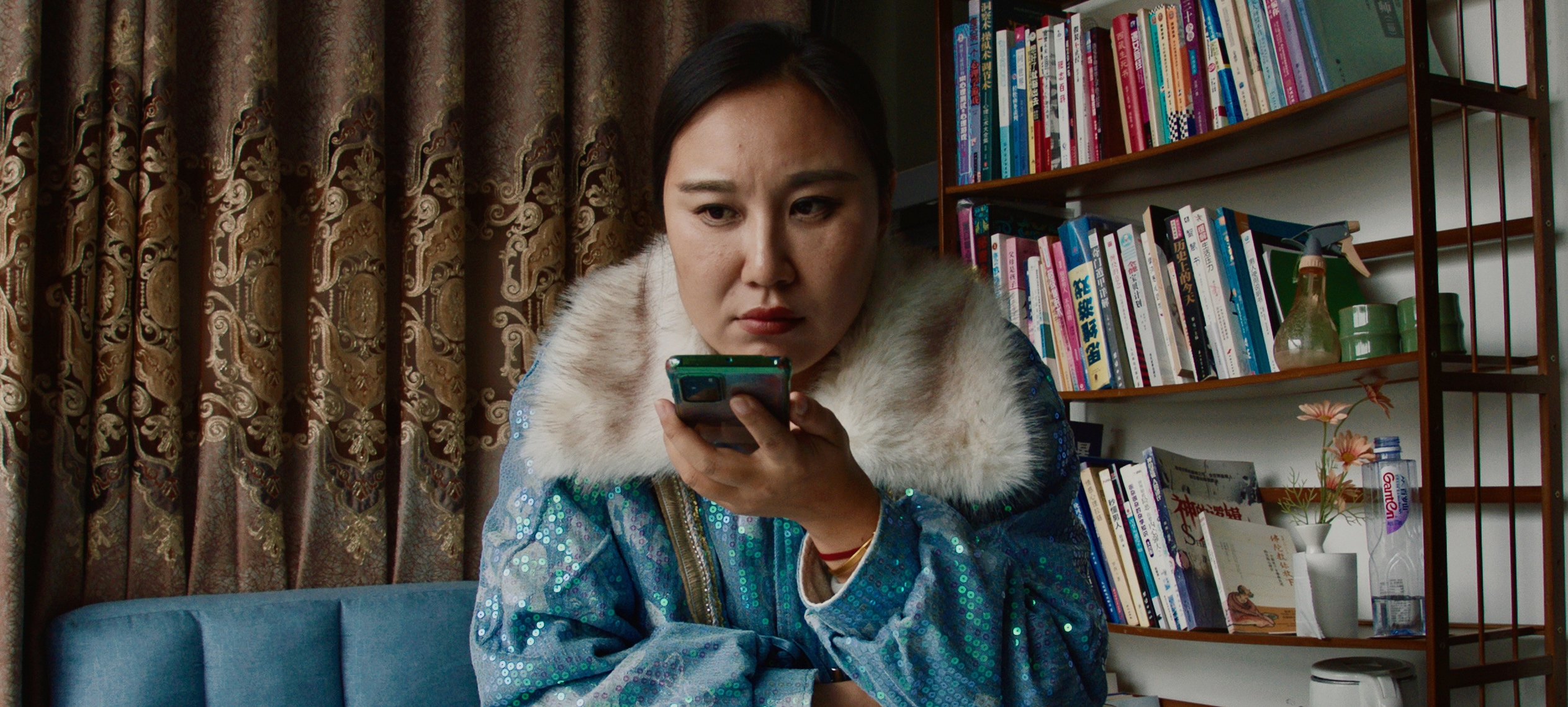 A person in a blue sparkly jacket with fur collar holds a mobile phone. they are seated at a table in front of a brocade curatin and a shelf of books.