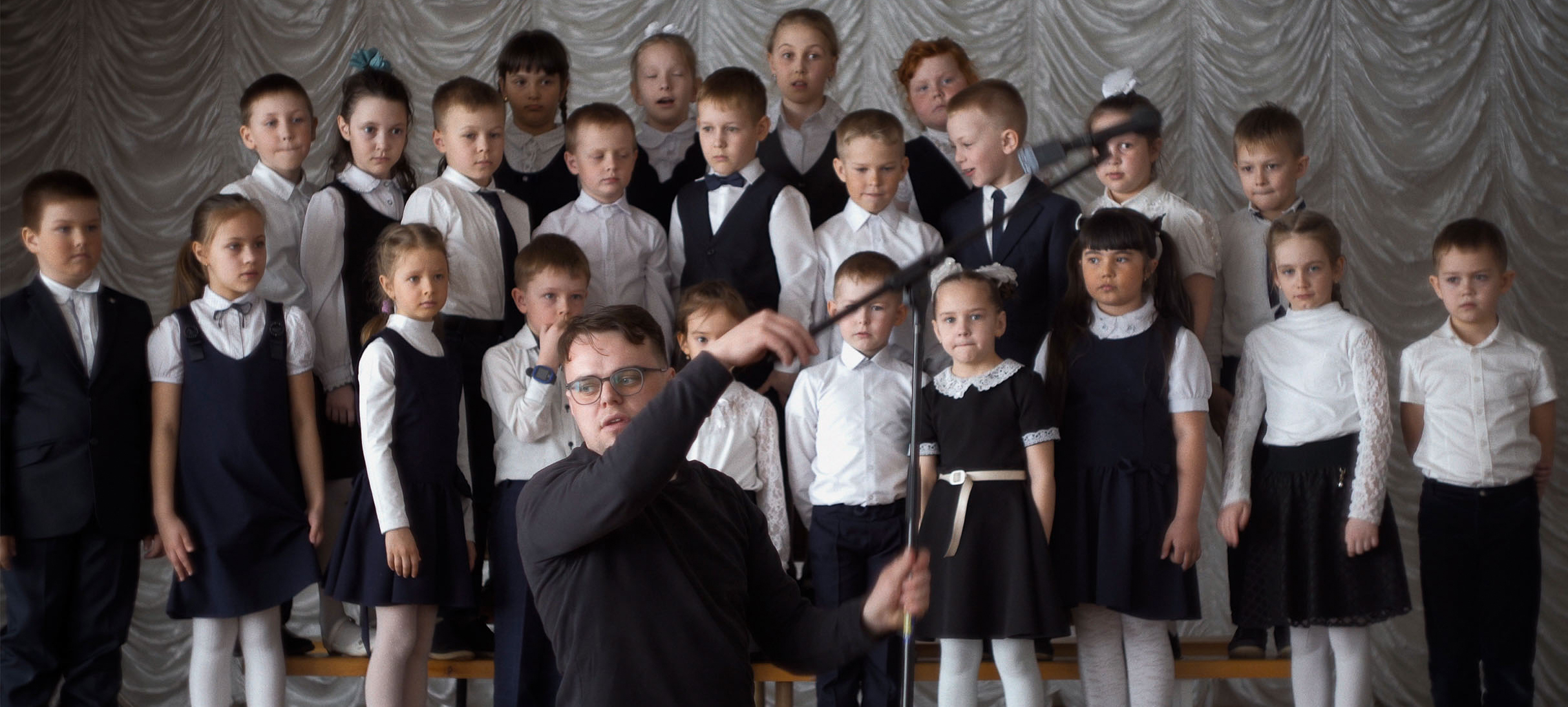 A conductor adjusts a mic in front of a group of children dressed in a black and white.