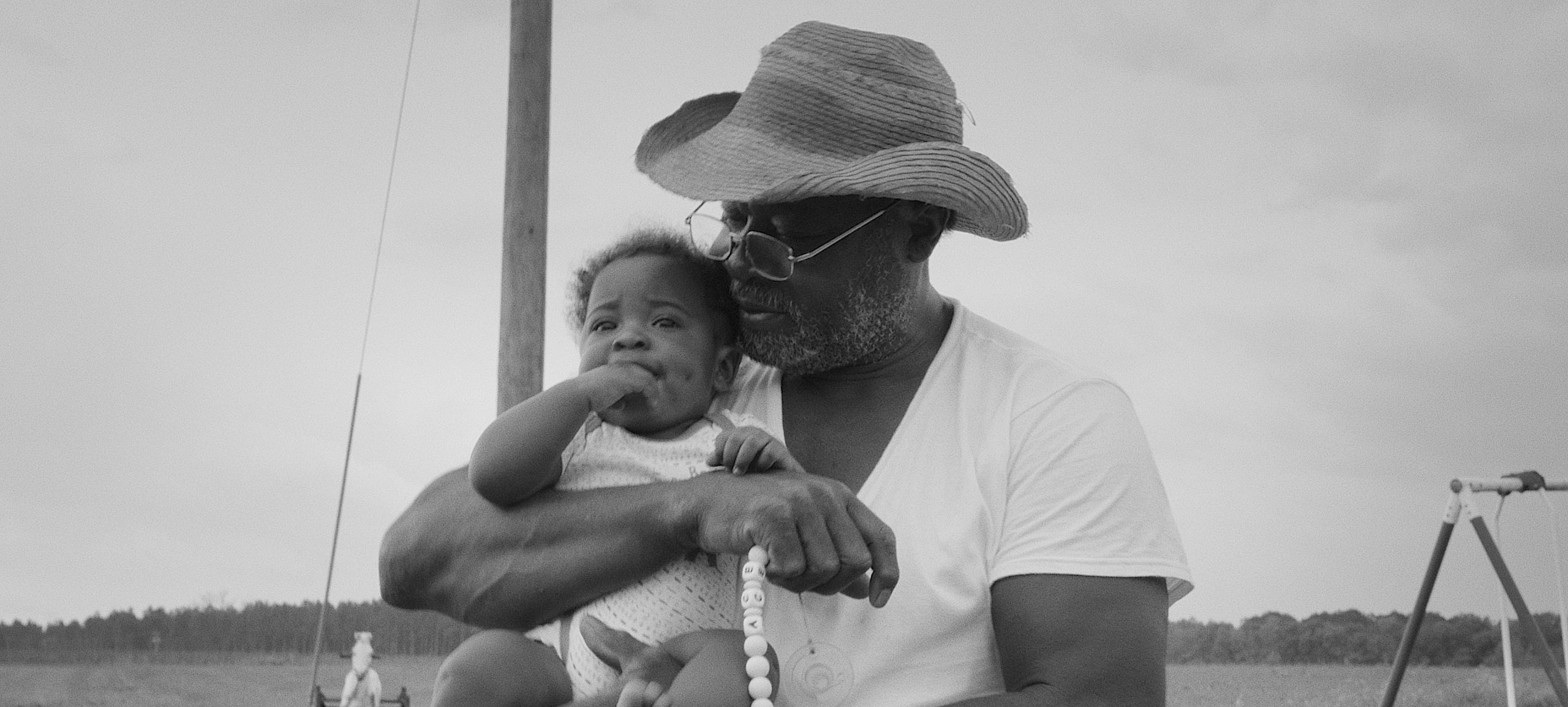 A black-and-white image of a person in a straw cowboy hat and white tee shirt, holding a baby in their muscled arms.
