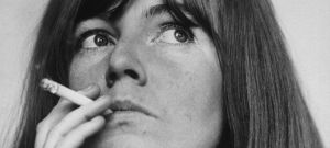 A black-and-white image of a person with long straight hair, smoking a cigarette. They are looking to the left of the camera.