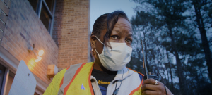 A person wearing a face mask and a safety vest stands in front of a brick building and looks toward the left with eyebrows furrowed.