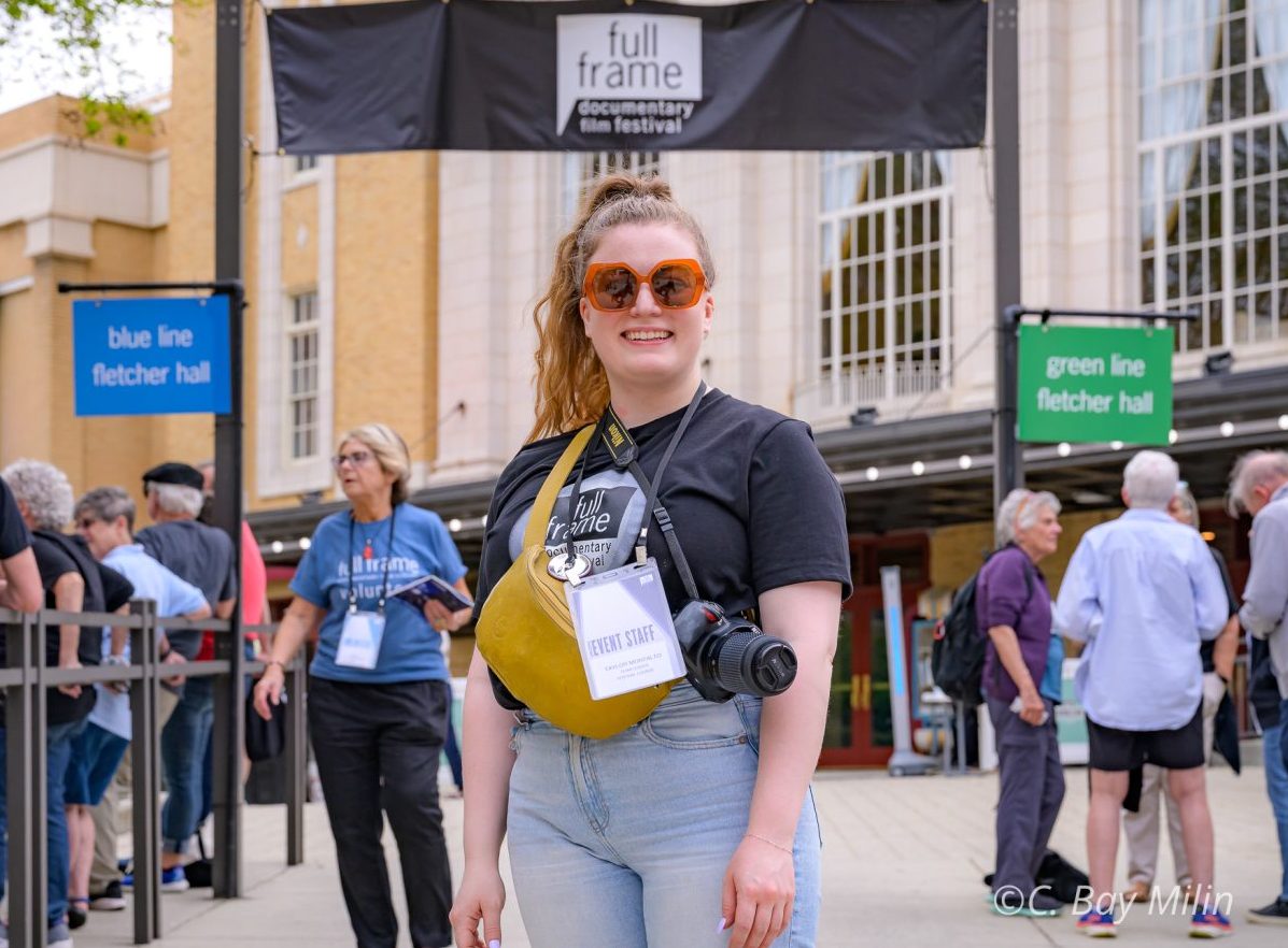 Volunteer stands in sunglasses with a camera in front of the Full Frame banner at festival