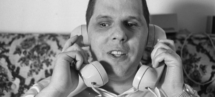 A man sitting on an upholstered sofa holds telephone receivers up to his mouth