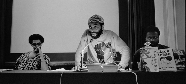 A man stands at a microphone, delivering comments. TYo his left, a seated man is surrounded by children's picture books on display.