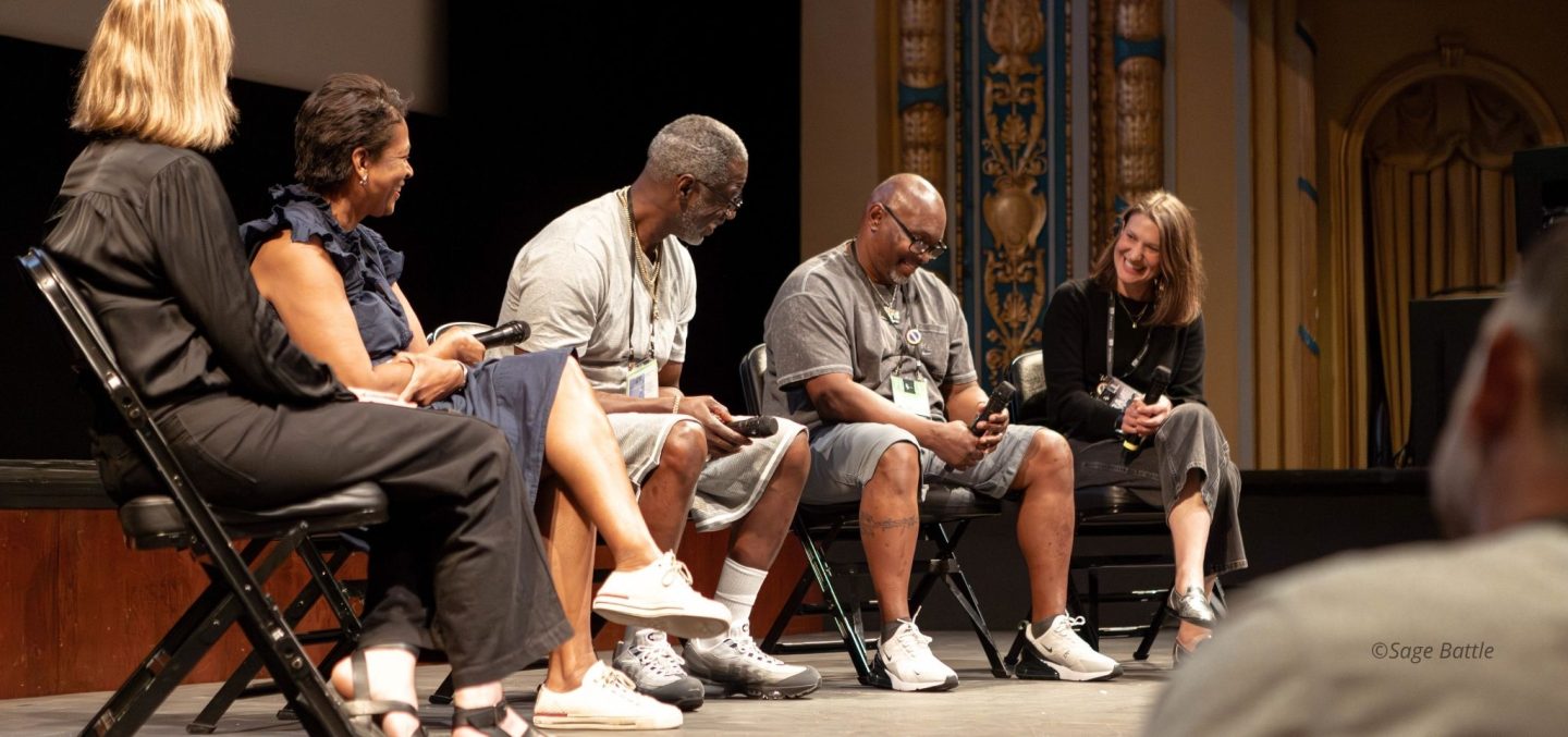 Film team from WHEN A WITNESS RECANTS onstage with story participants and festival co-director. They are seated in folding chairs onstage, laughing.